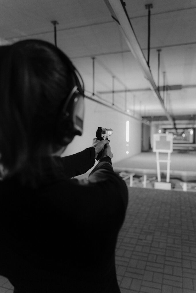Black and white image of a woman shooting at an indoor range with headphones and pistol.